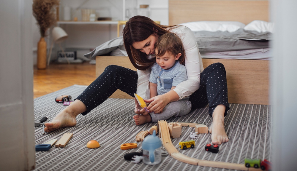 mama care sta pe podea cu bebelusul in brate langa jucarii de lemn