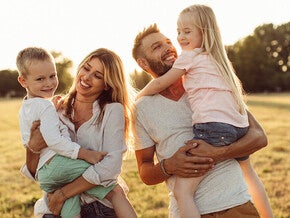 Photograph of smiling parents holding young son and daughter in sunlit grassy field