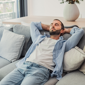 Photo of a relaxed man reclining on a gray sofa, eyes closed, hands behind head.