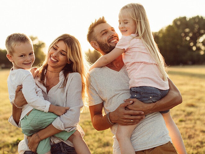 Photograph of smiling parents holding young son and daughter in sunlit grassy field