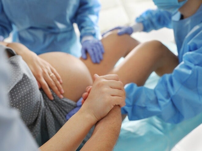 Fotografie: femeie în travaliu asistată de echipă medicală; partener îi ține mâna