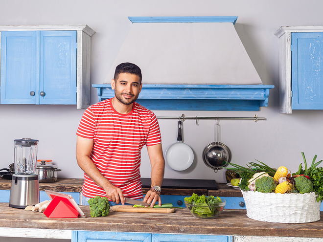 Photo of smiling man in red-striped shirt chopping vegetables in blue kitchen