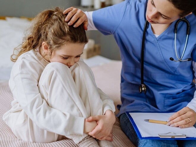 Medic îngrijorător consultă un copil pe pat, ținând un clipboard.