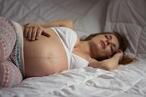 Femeie însărcinată întinsă în pat, cu mâna pe abdomen, odihnindu-se din cauza disconfortului asociat sarcinii.