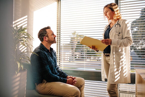 Doctor in white coat with clipboard speaking to seated male patient in sunlit clinic